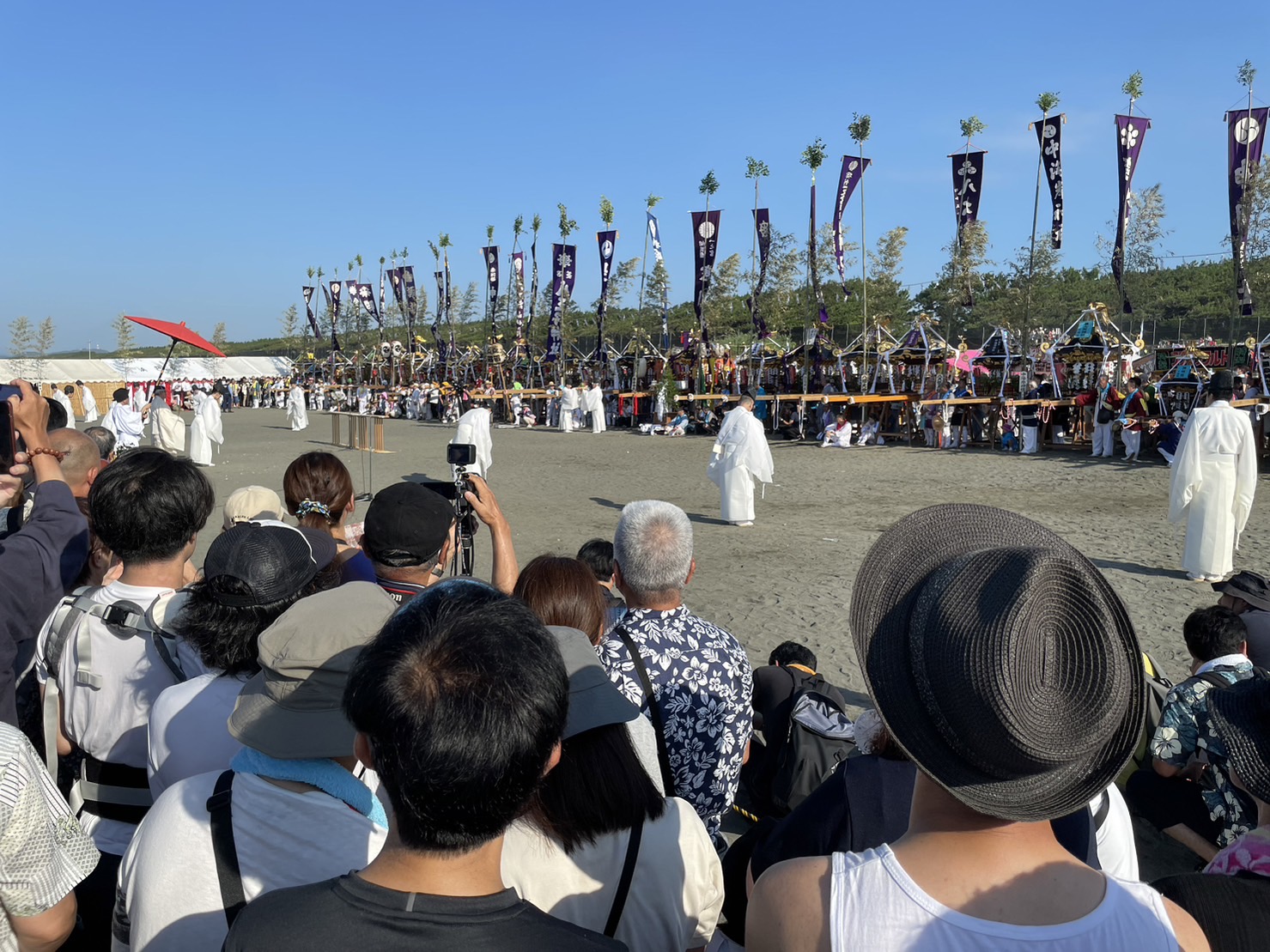 浜降祭の浜辺で、白装束の神職が儀式を行う様子。背後には多数の神輿と紫の旗が並び、朝日を浴びる観客が見守っている。
