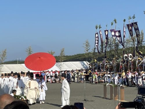 浜降祭の儀式で、赤い和傘を差した神職の行列が進む様子。背景には神輿と祭旗が並び、青空の下で多くの参拝者が見守っている。