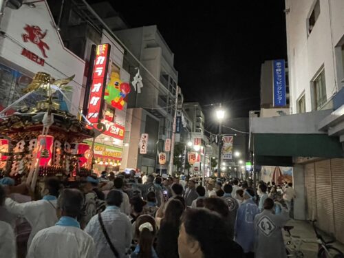浜降祭の夜、茅ヶ崎駅北口の商店街を神輿が進む様子。担ぎ手たちが法被姿で列をなし、周囲の看板や提灯が明るく照らす中、観客が見守っている。